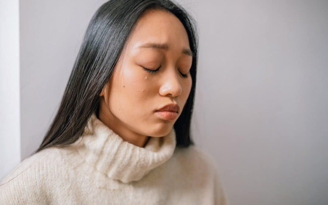 Asian woman with long straight hair and wearing a turtleneck cries.