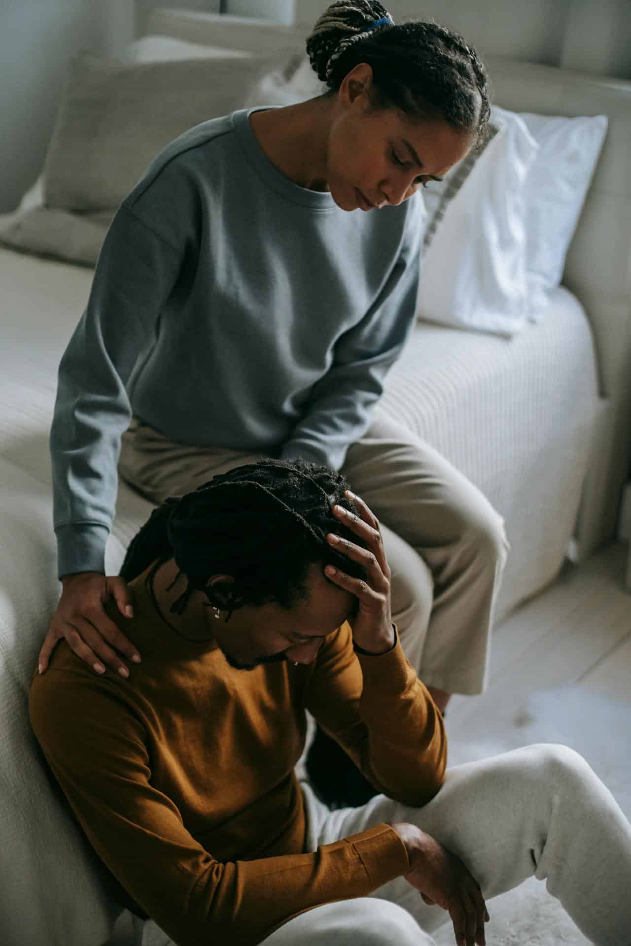 A woman sits on the edge of a bed, gently comforting a man sitting on the floor with his head in his hands. The man appears distressed, while the woman offers support with her hand on his shoulder.