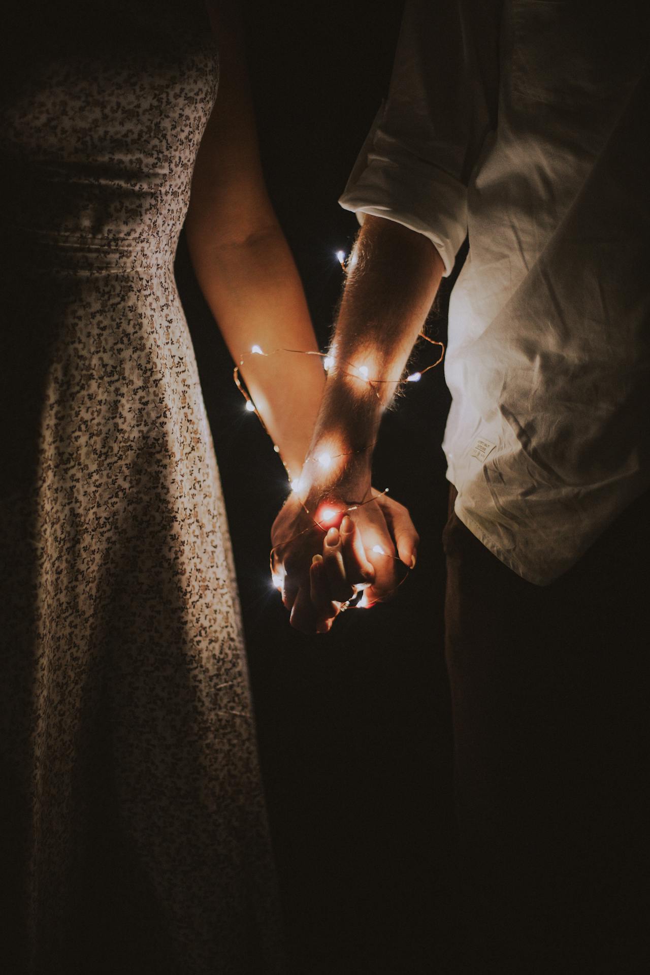 Half of the torso man dressed in a white long-sleeve shirt and woman in a flowery dress, standing holding hands with twinkly lights wrapped around their arms. 