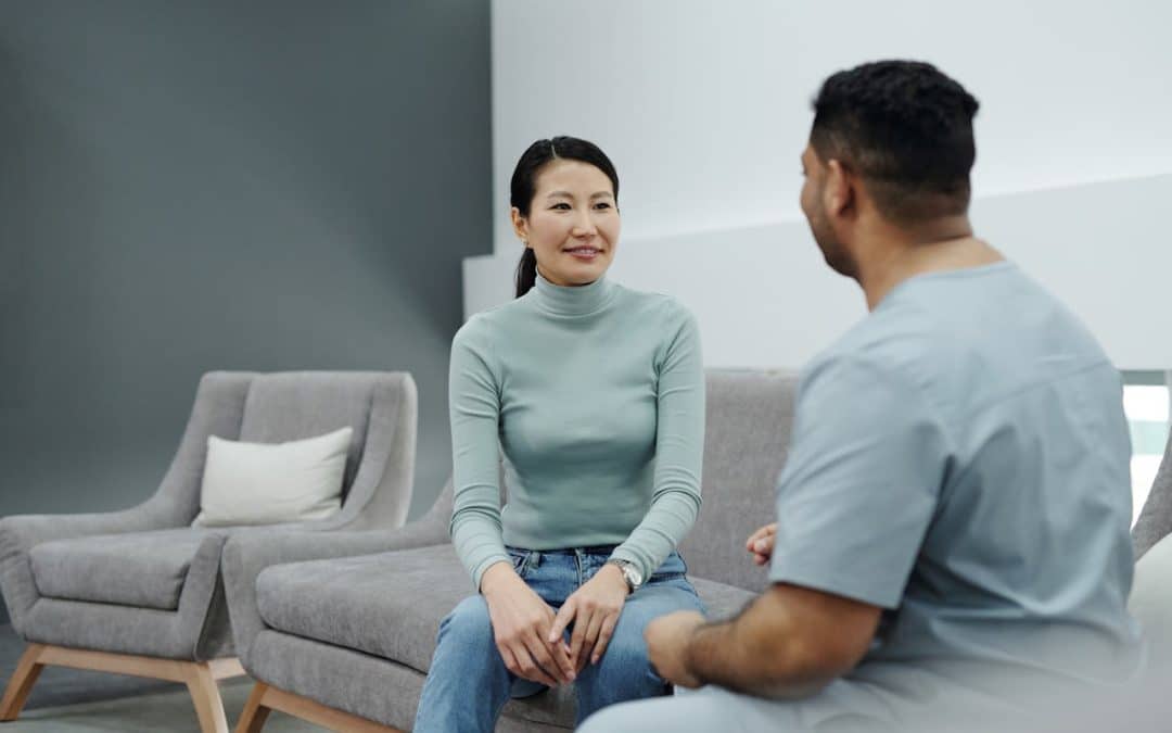 An asian woman sitting on a couch talks to a man across from her. She wears a light blue turtleneck, and he is wearing a grey t-shirt.