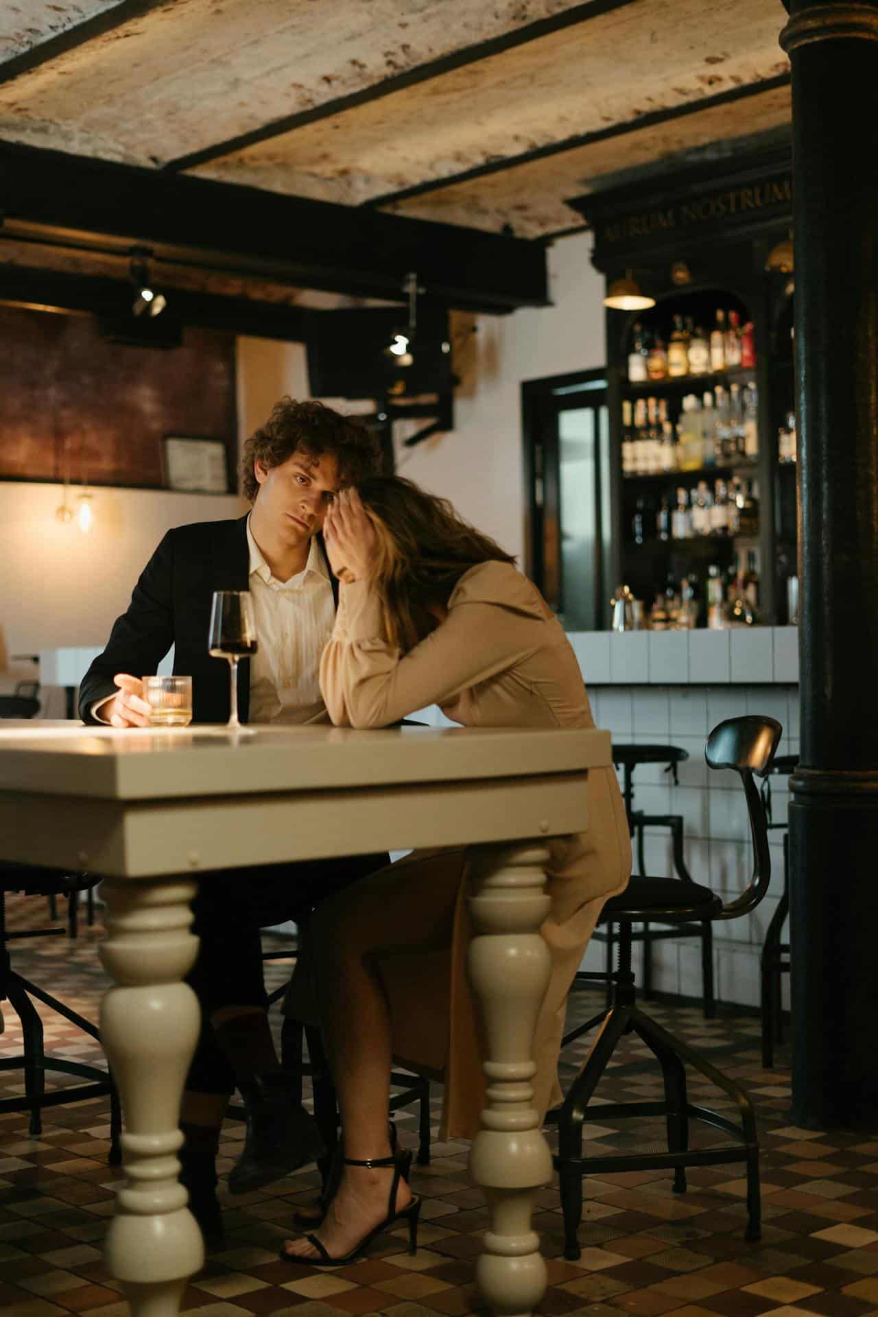 A man and a woman are sitting at a table in a dimly lit bar or restaurant. The man, dressed in a suit, looks straight ahead while holding a drink. The woman, wearing a beige dress, leans on the table with her head in her hands, appearing distressed.