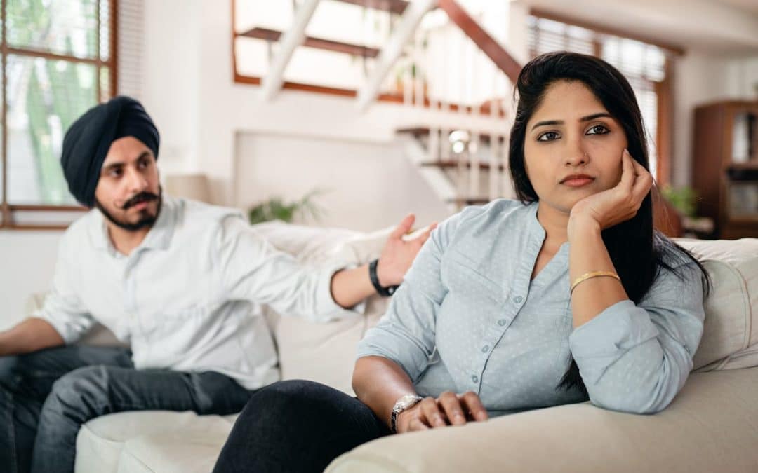 A man and a woman are sitting on a couch in a living room. The man, wearing a turban and a white shirt, is reaching out to the woman, who looks away with a thoughtful or upset expression.