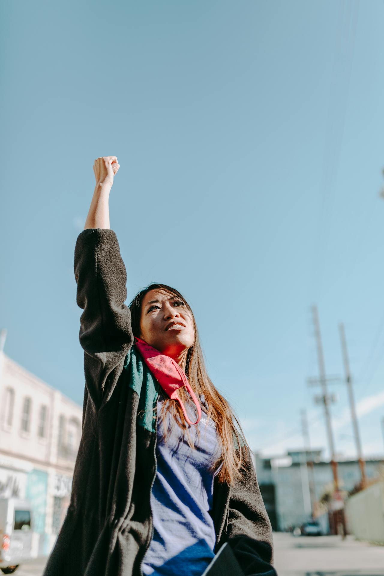 An Asian Woman wearing a black jacket and red mask raises her hand in the air.