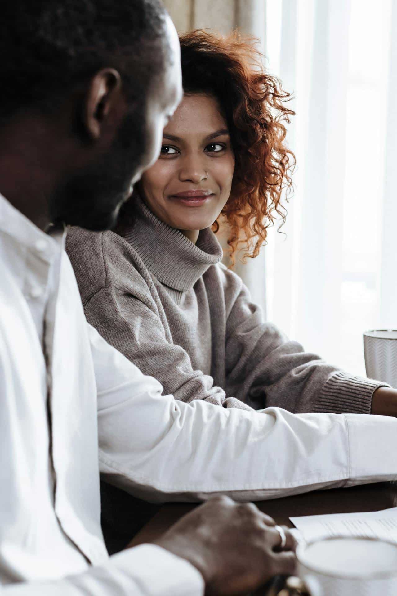 A young black woman with brown and curly hair, wearing a grey turtleneck, looks at a black man sitting next to her, smiling.