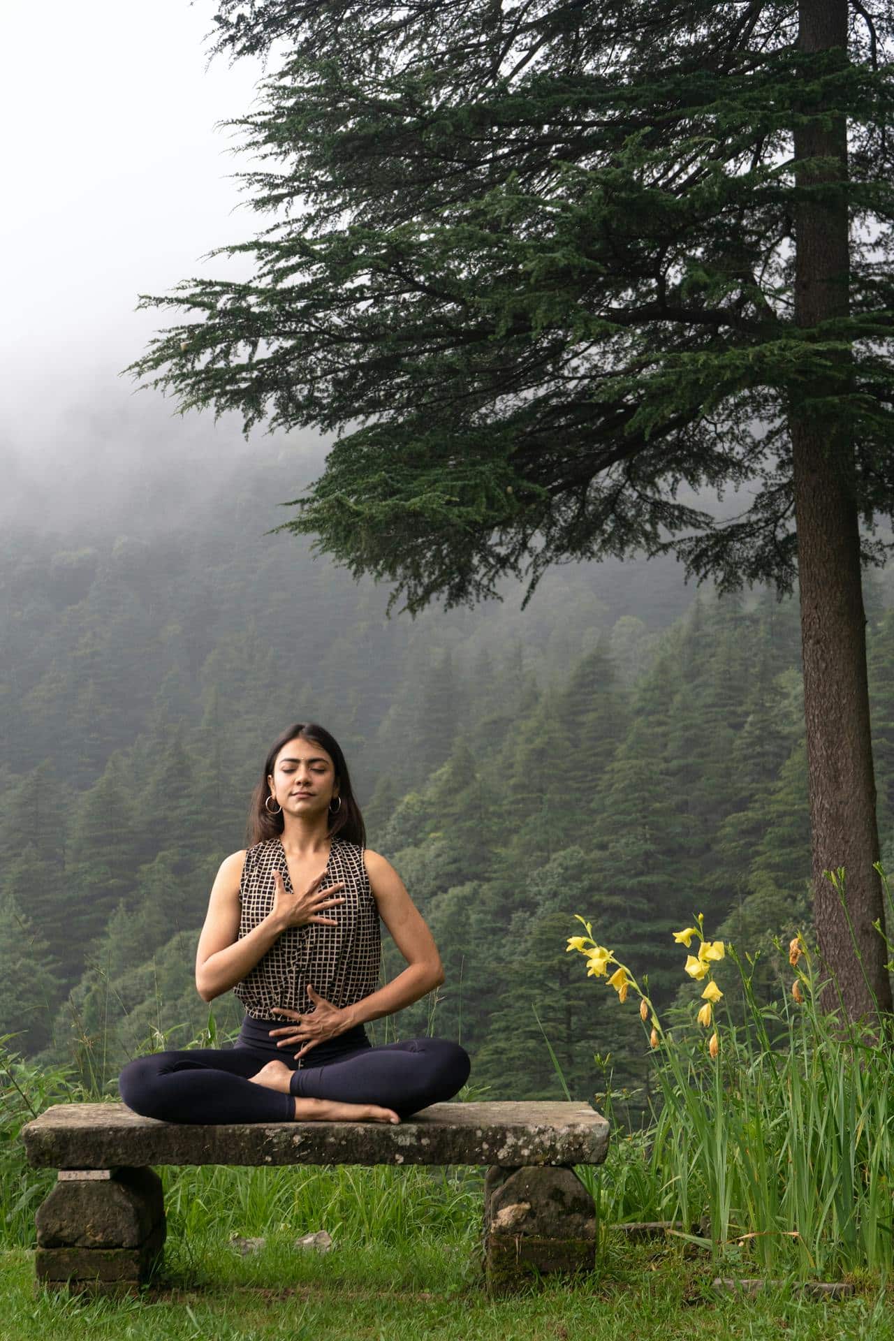 A woman with long dark hair sits cross-legged on a wooden bench, practicing mindfulness meditation. A forest scenery is behind her. 