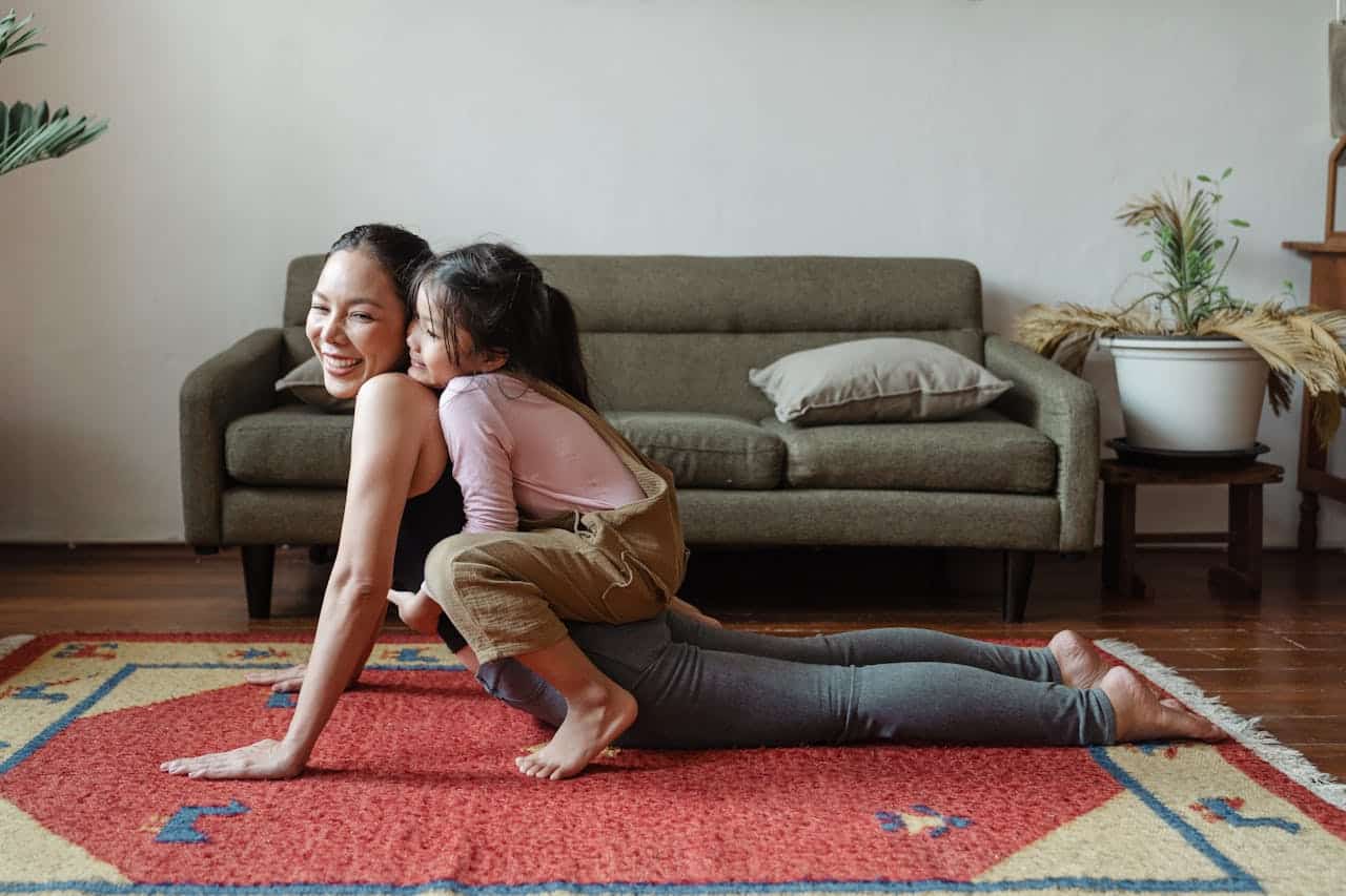 An Asian woman laughs while doing a yoga pose, while a young Asian child playfully hugs her back.