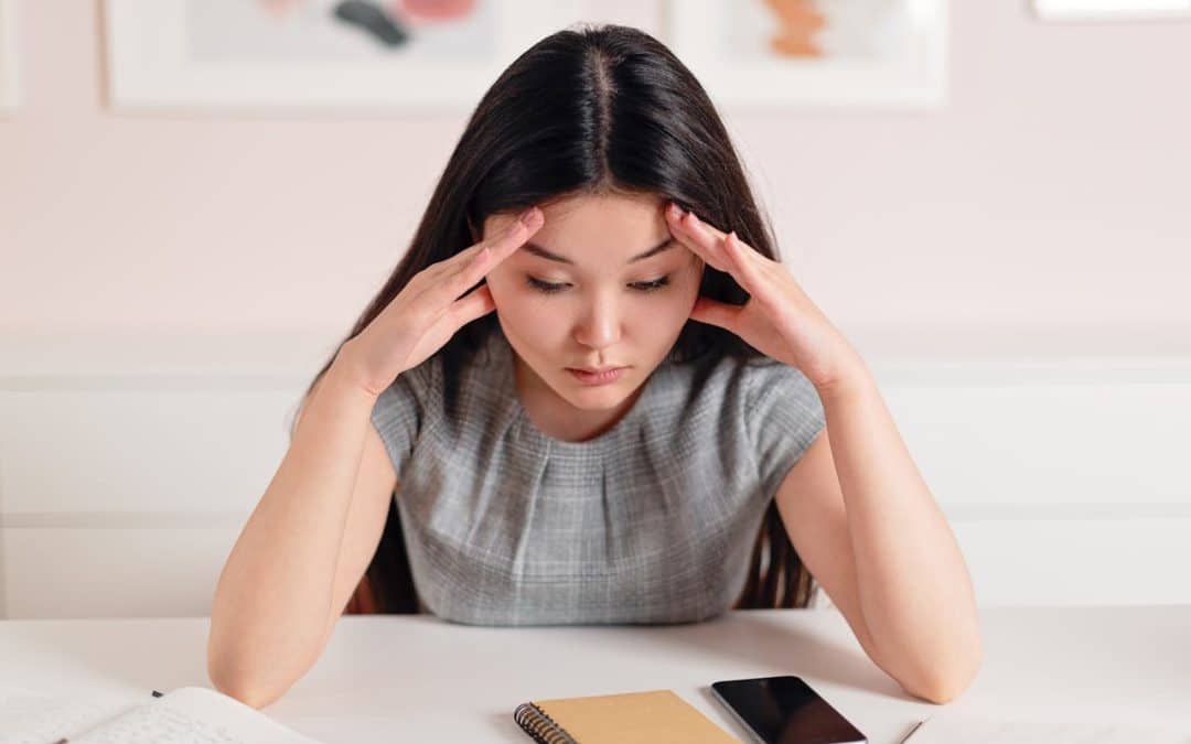 A young Asian woman with long straight black hair sits on a table with her hands on her forehead, looking down at her desk.