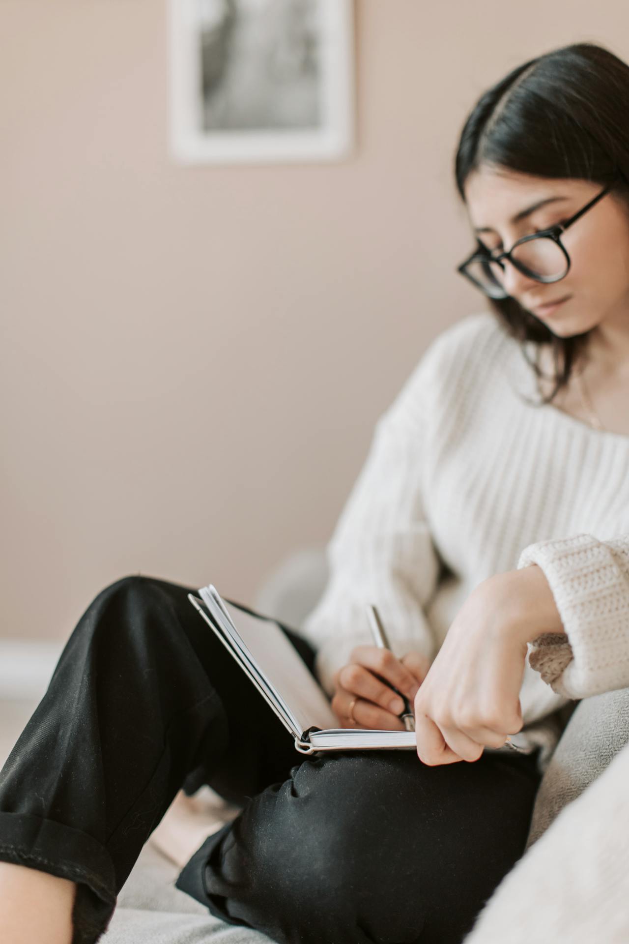 A woman with dark hair, wearing a white sweater and black pants and dark glasses, sits on her couch writing in her notebook.