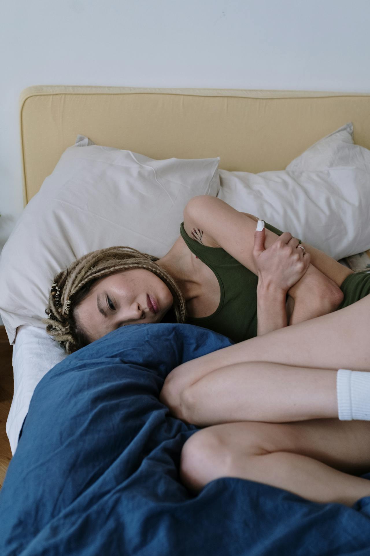 A woman lies curled up in bed, hugging herself with a somber expression, surrounded by pillows and blankets.