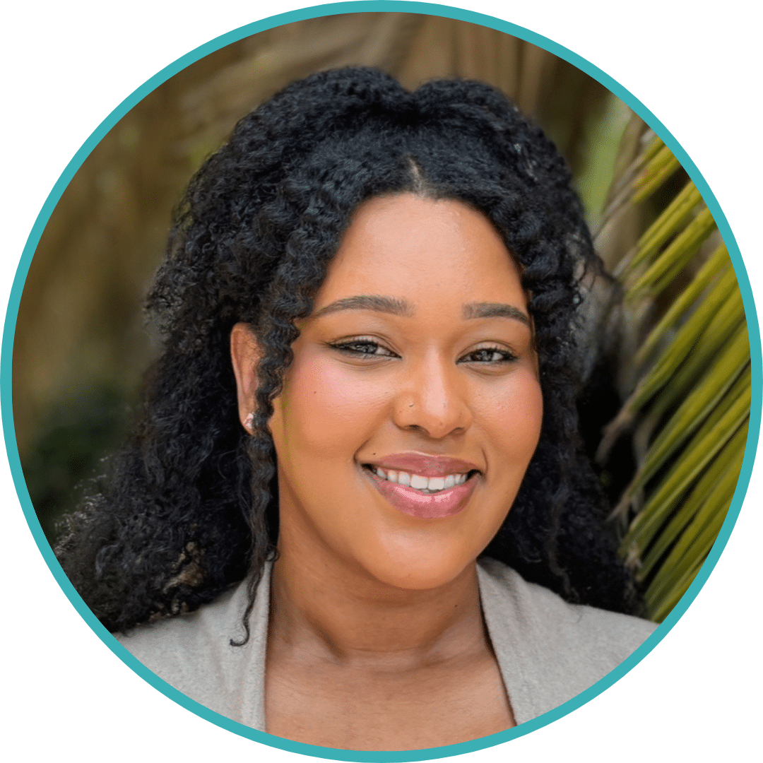 Headshot of Kila Johnson, a woman with a medium complexion and dark curly hair tied half back. She is wearing a beige cardigan over a blue top, and smiling at the camera. Palm trees and other foliage are visible in the background.