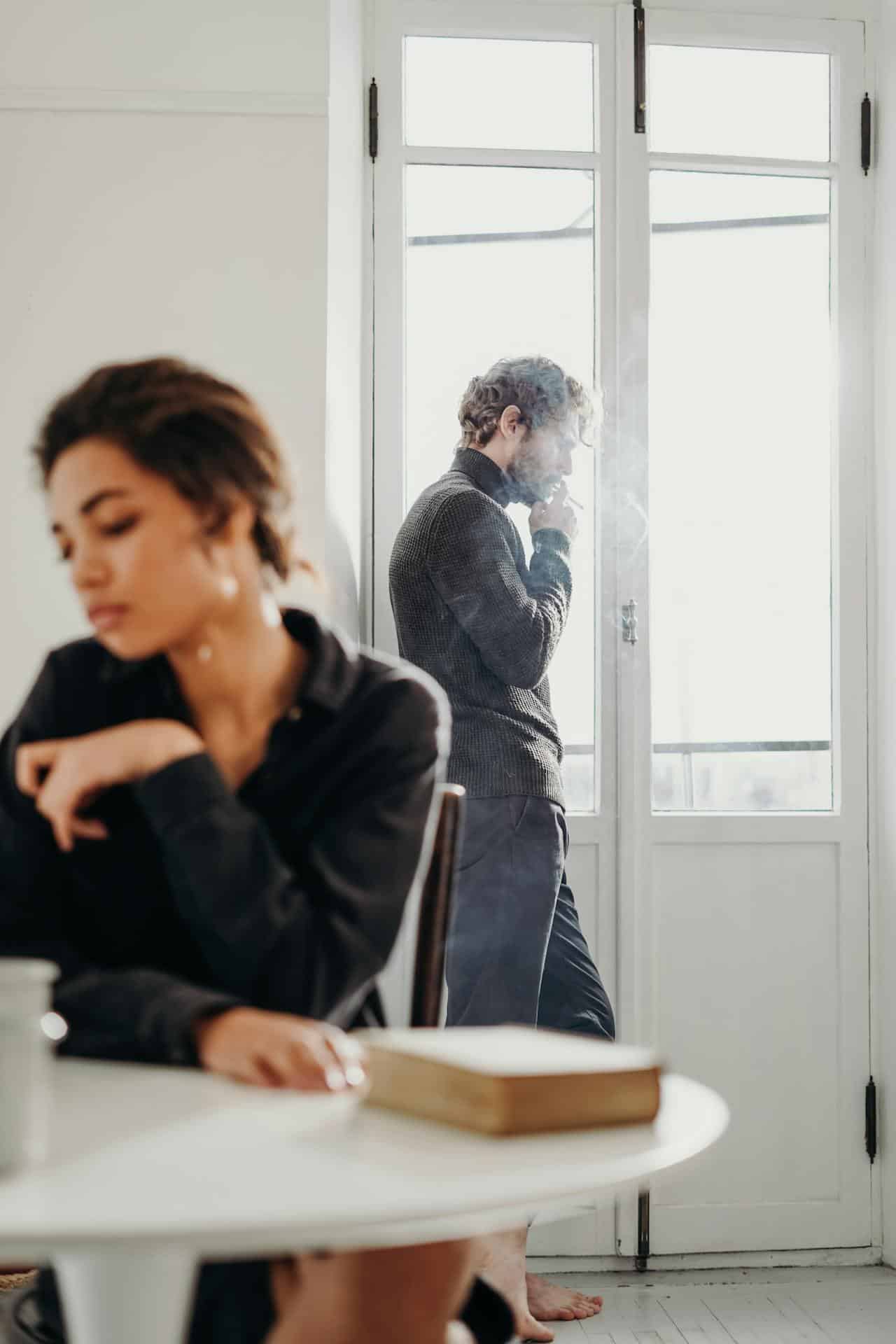 Woman sitting while man stands at the window, smoking.