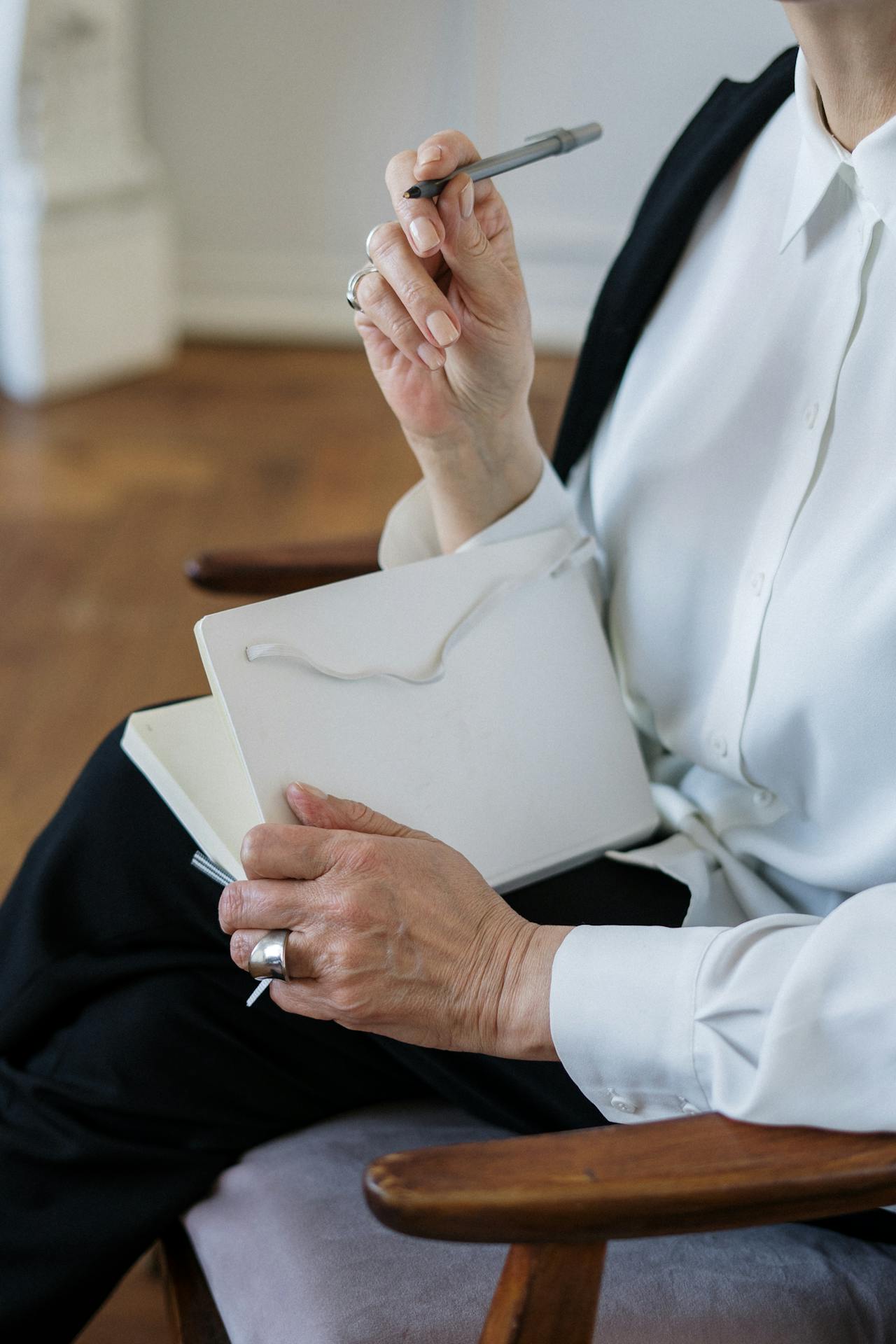 A woman in a white blouse holds a white notebook and a pen as she contemplates what to write.