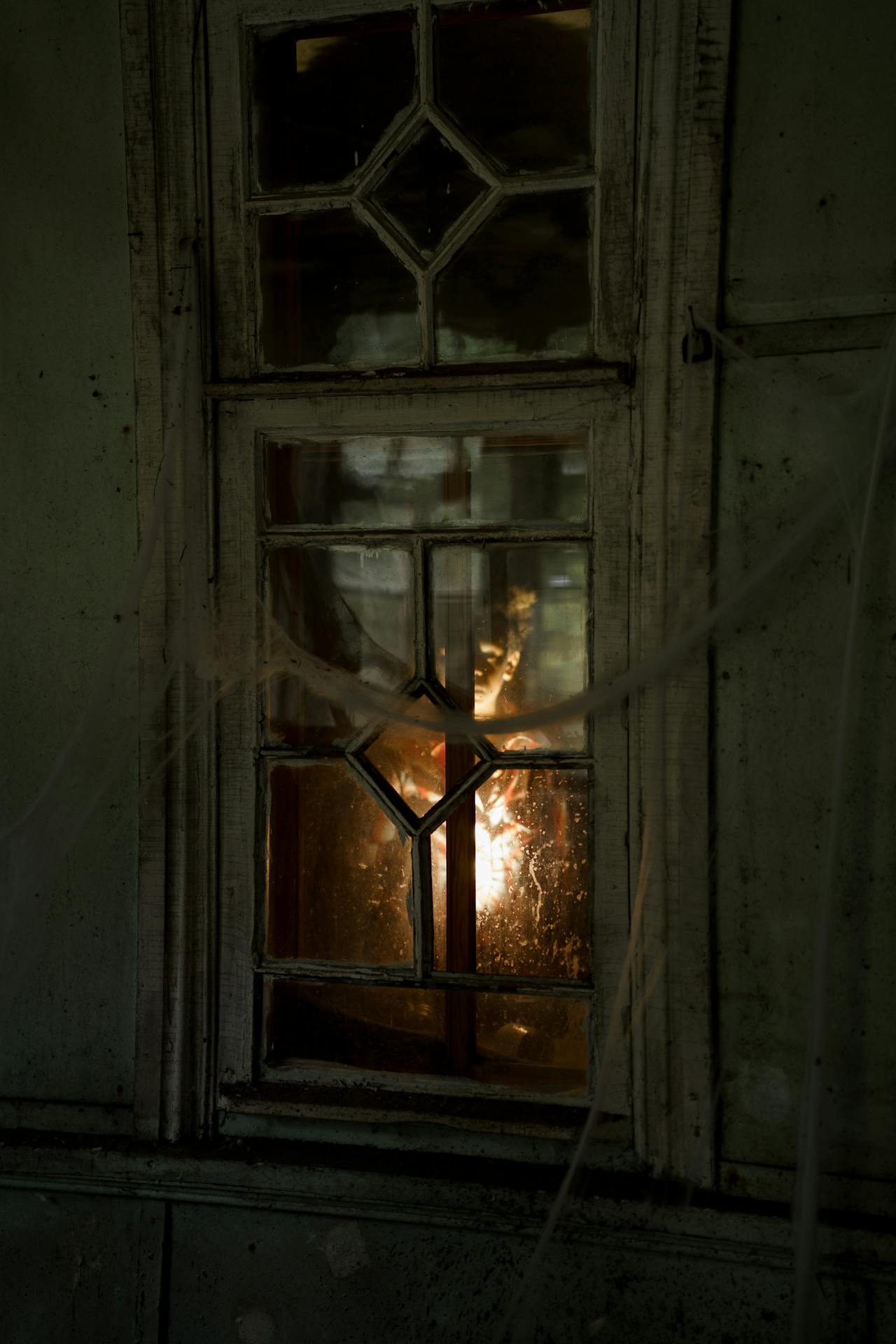 A figure holds a candle on the other side of a spooky looking window.