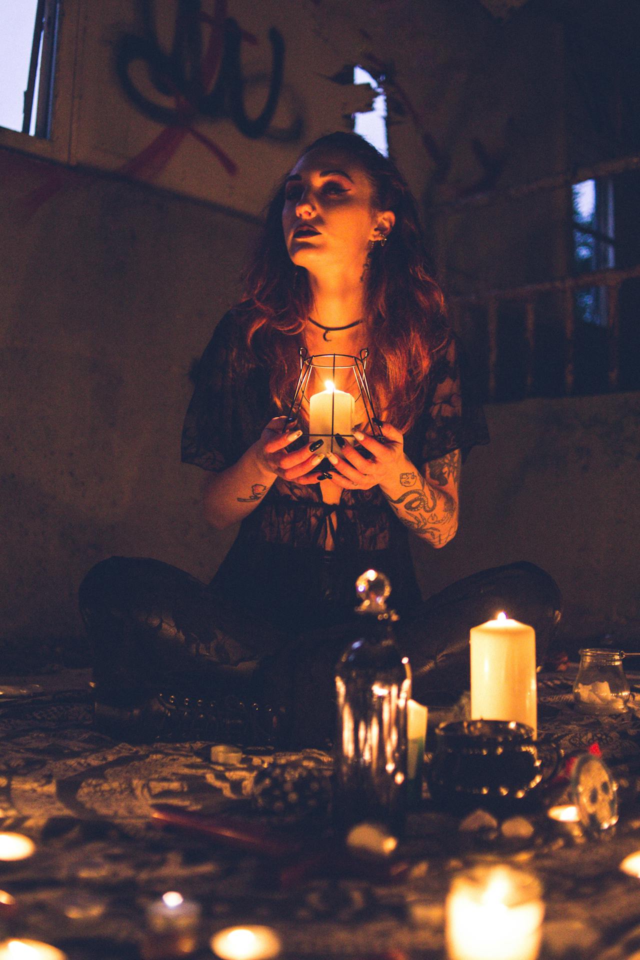 A witchy woman sitting on a tapestry on the floor surrounded by candles and occult objects