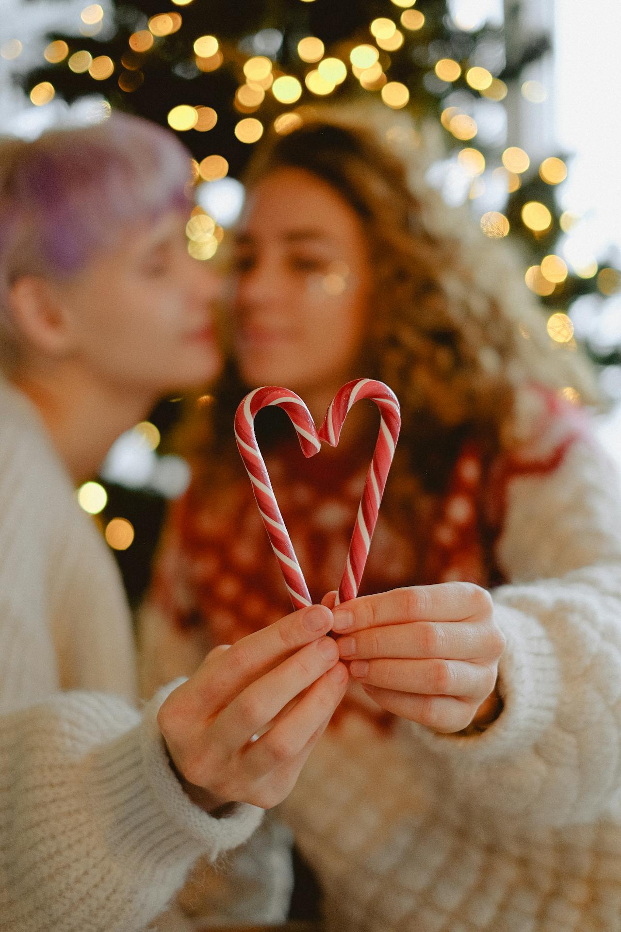A lesbian couple stand in front of a Christmas tree while holding candy canes in the shape of  a heart
