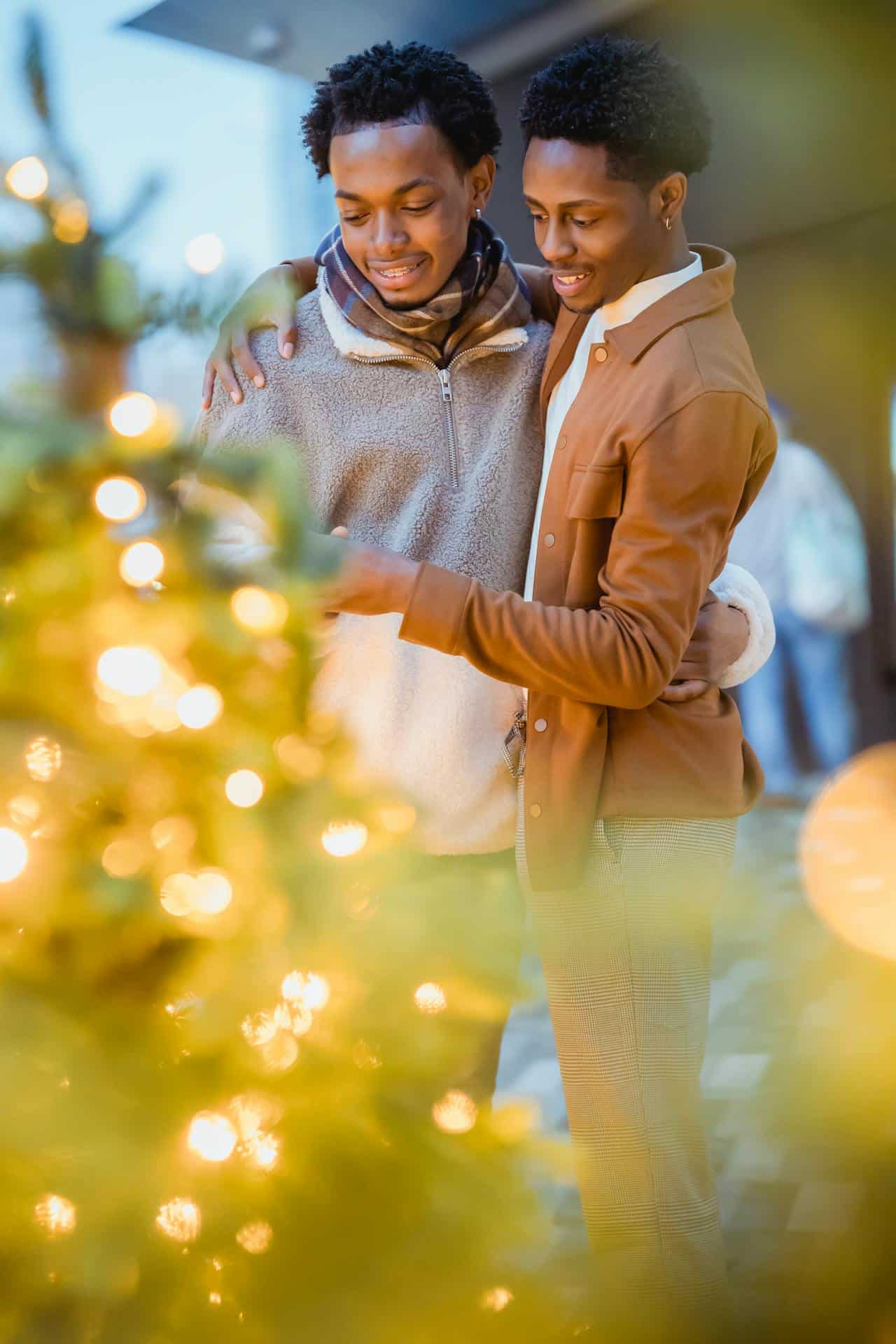 A black gay couple stand with their arms around each other in front of a Christmas tree