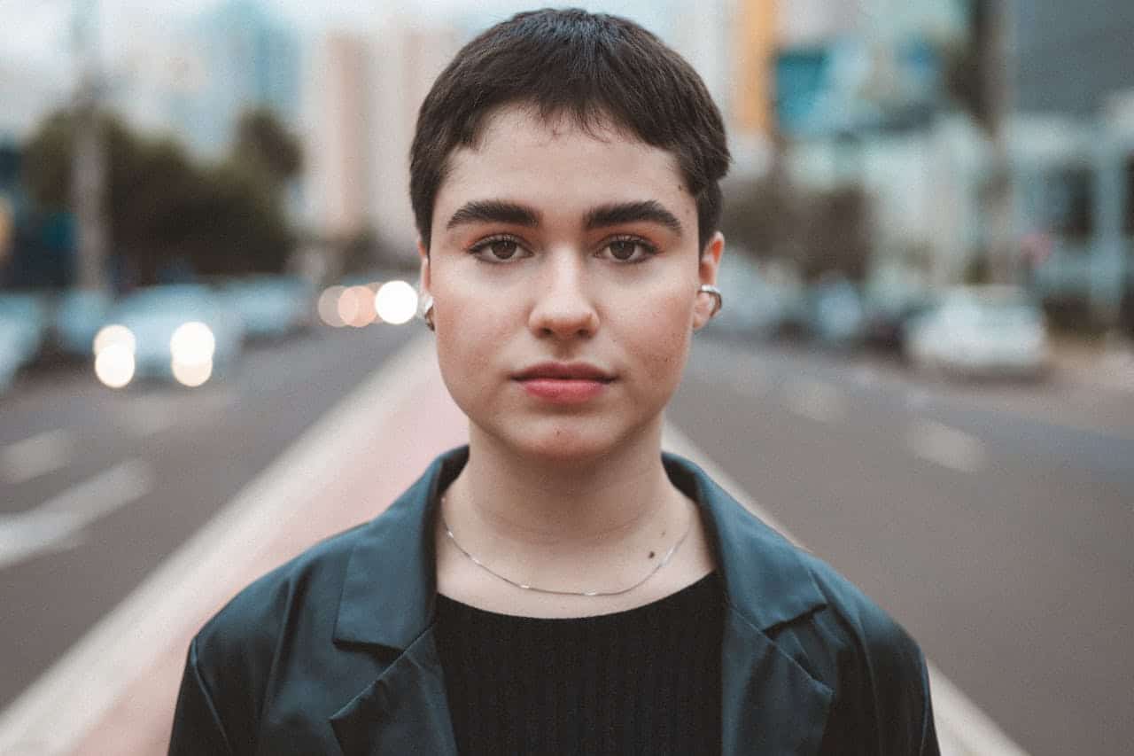 An androgynous person with short hair and wearing a leather jacket stands in the street