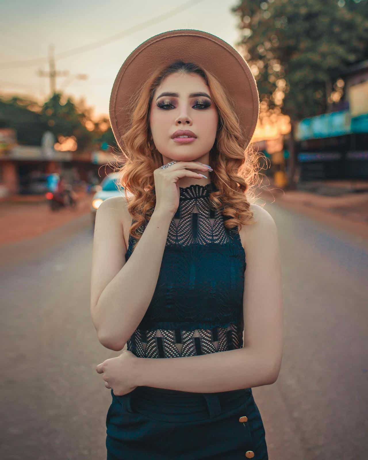 A Latine woman in a black dress and tan colored hat poses at sunset in teh street